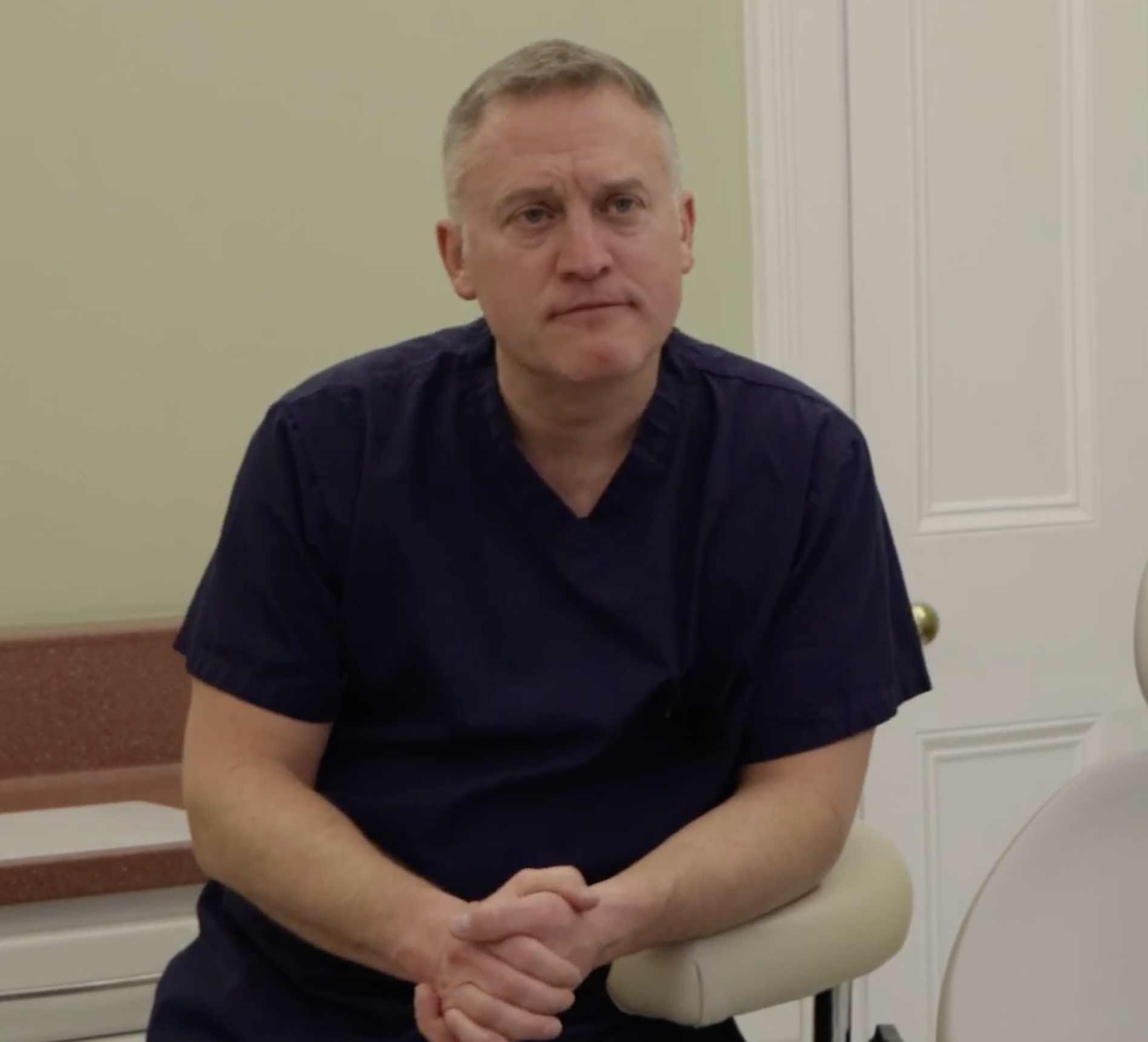 a man in a blue scrub is sitting in a dental chair with his hands folded .