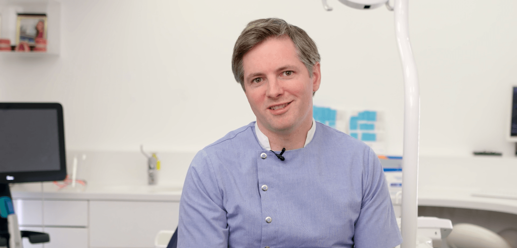 A smiling man in a light blue scrub top in a dental office.
