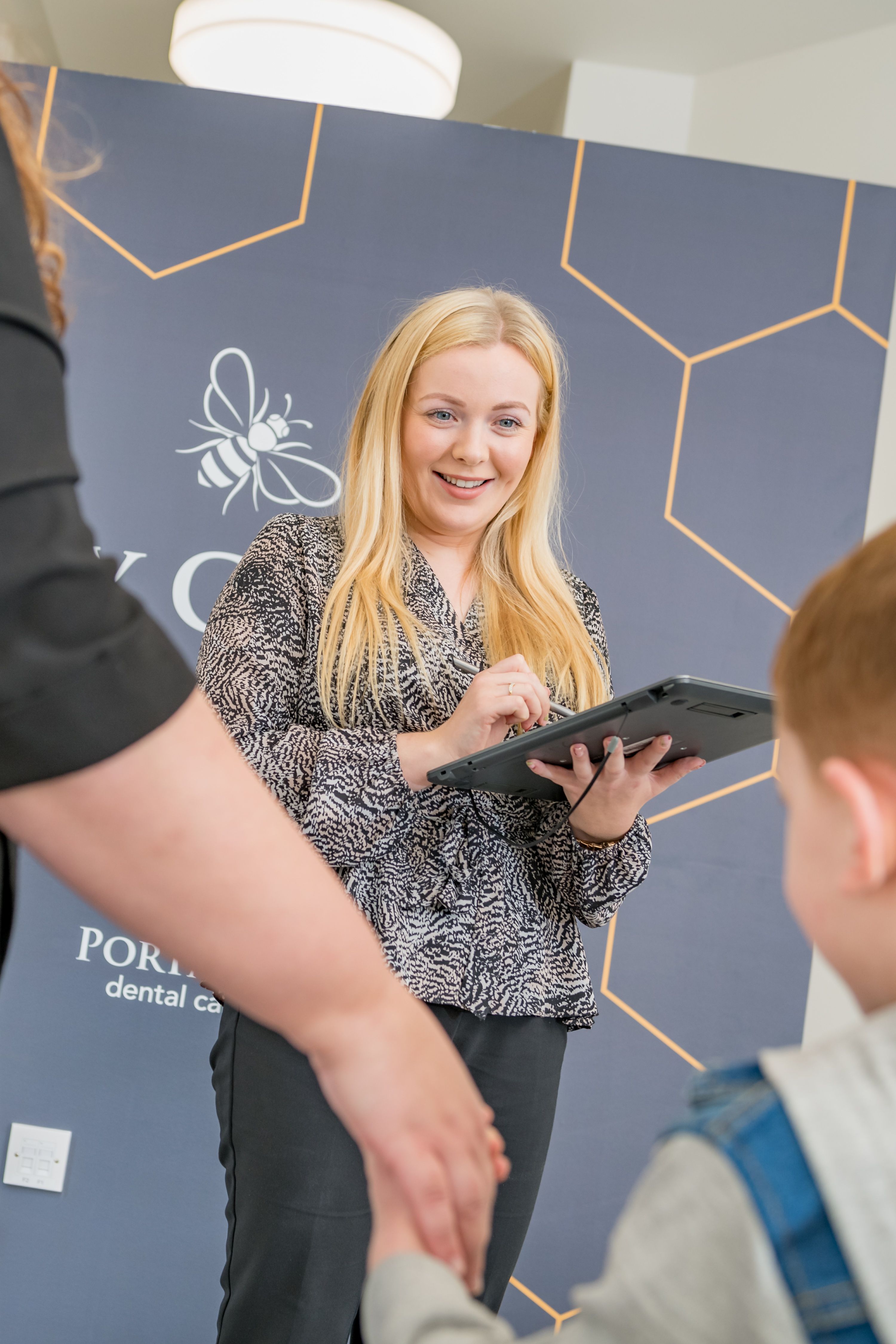 A smiling blonde woman holding a tablet looks at patients in a dental clinic.